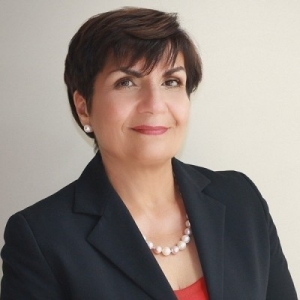 A woman with short, dark hair and a pearl necklace stands in front of a beige background