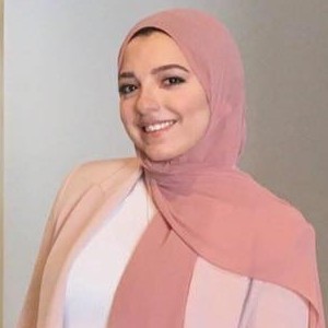 A woman in a pink hijab stands in front of a plain background