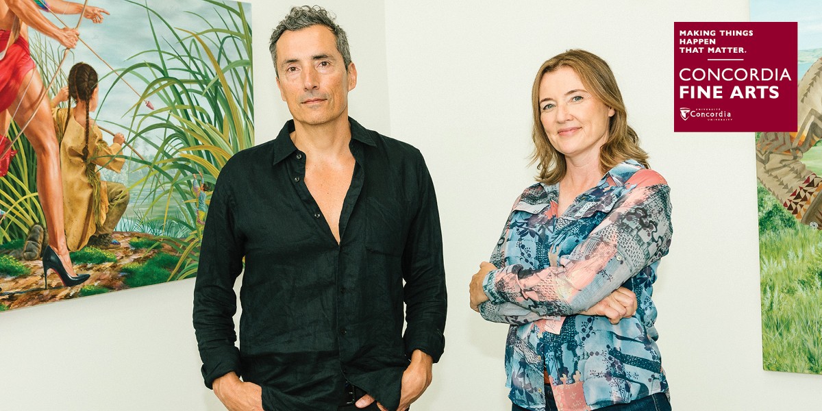 Kent Monkman, wearing a black button down, and Gisèle Gordon, wearing a blue patterned blouse, stand in front of one of his paintings. 