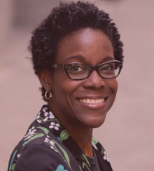 Portrait of a smiling woman in glasses, dressed in a structured floral jacket against a pink background.
