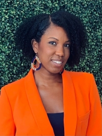 A woman with dark, curly hair, wearing a black top, orange blazer and gold earrings smiles at the camera