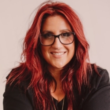 Portrait of a smiling woman in glasses, dressed in a structured floral jacket against a pink background.