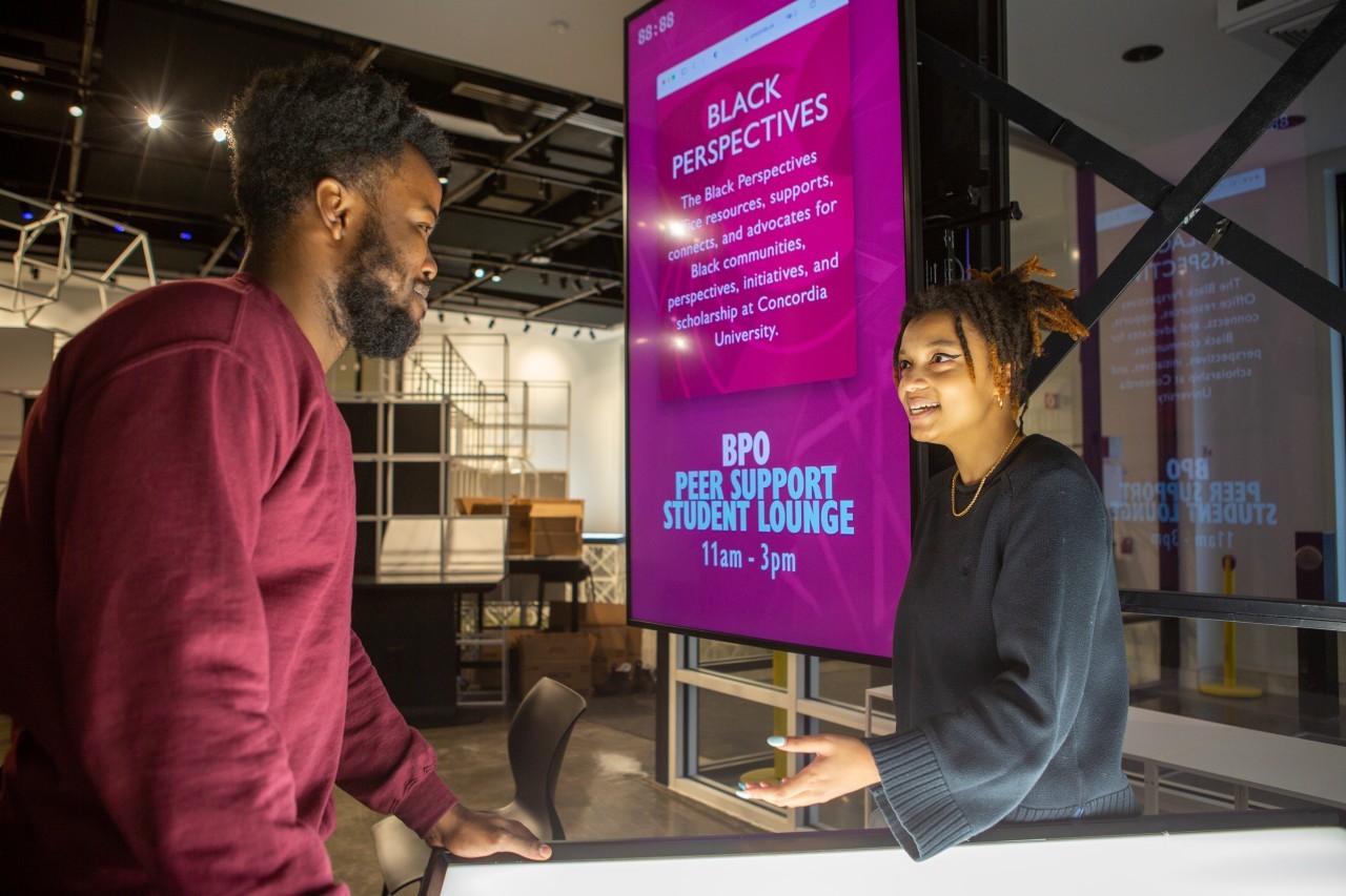 A Black woman standing beside an advertisement for the Black Perspectives Office, speaking to a Black man.