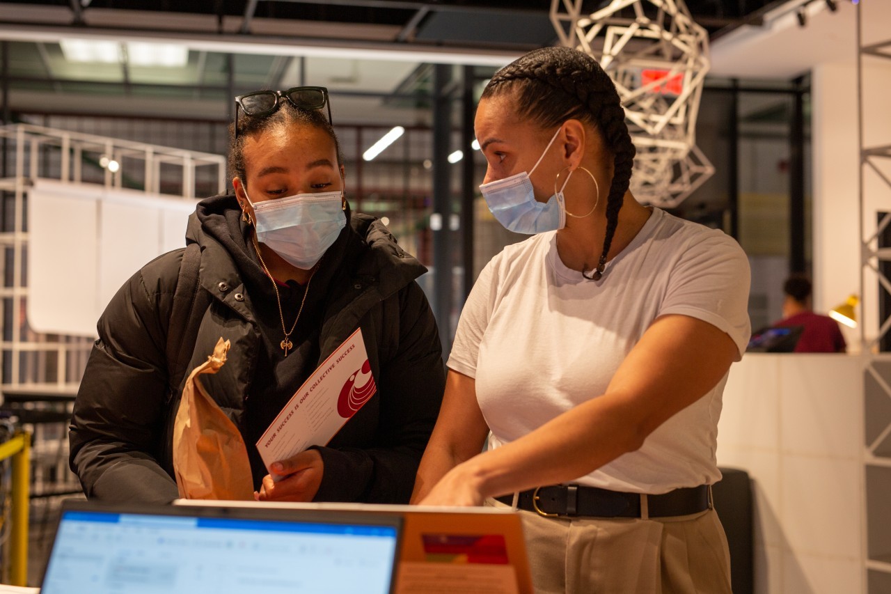 Two Black women wearing blue face masks talking in front of a computer screen