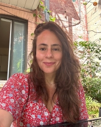 A person with long, wavy brown hair sits outside in a sunny courtyard, wearing a red floral top with plants and a brick wall behind them.