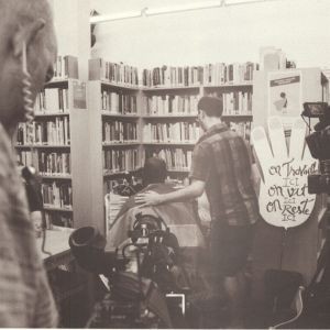 Black and white photo of a person touching another person's shoulder in a library, surrounded by video camera and a sign reading "On travail ici, on vit ici, on reste ici."