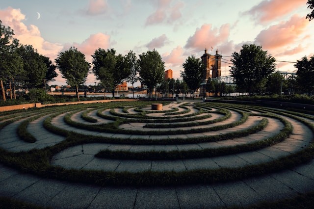 An outdoor labyrinth made of plants and stone paths, with trees and a sky visible in the background.