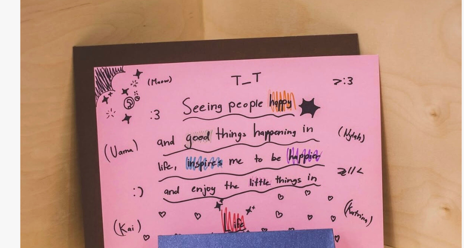 On a pink paper saying 'Seeing people happy and good things happening in life, inspires me to be happier and enjoy the little things in life' with different names on the sides as this was a collaboration. A blue paper leaning up against the bottom of it has one name on it.