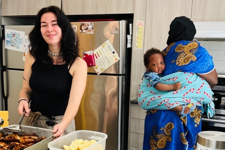 Two women preparing food in the kitchen. The woman at the stove is baby-wearing.