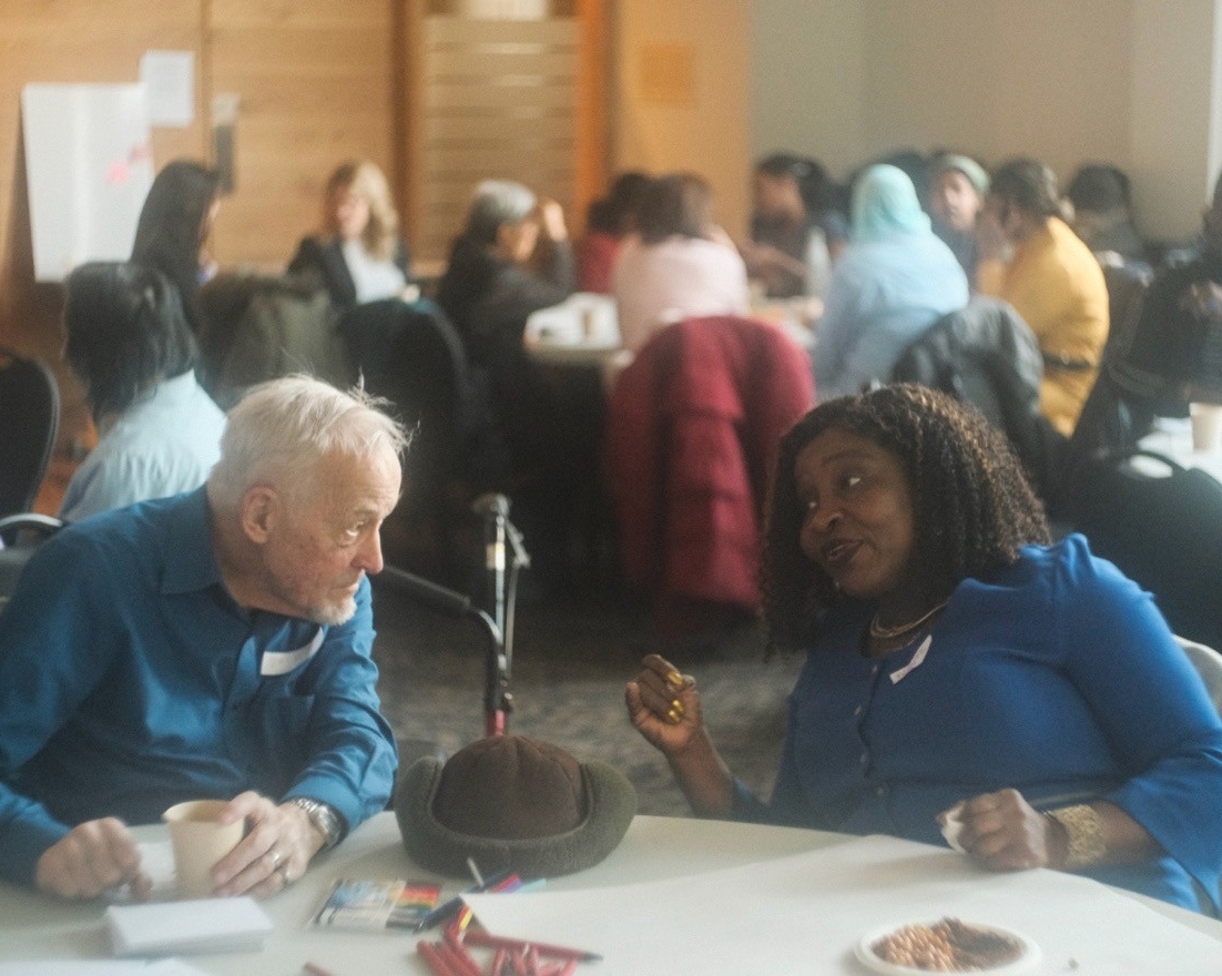 An older white man and an older black woman sit around a table with food leaning in closer to each other engaging in lively conversation.