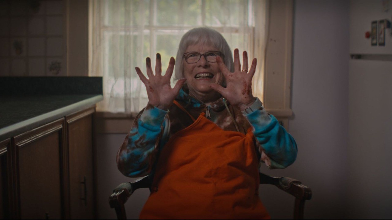 An older woman with white hair and glasses, wearing an orange apron, strikes a horror movie pose with her hands raised defensively palm out. Red specks on her hand are suggestive of splattered blood.