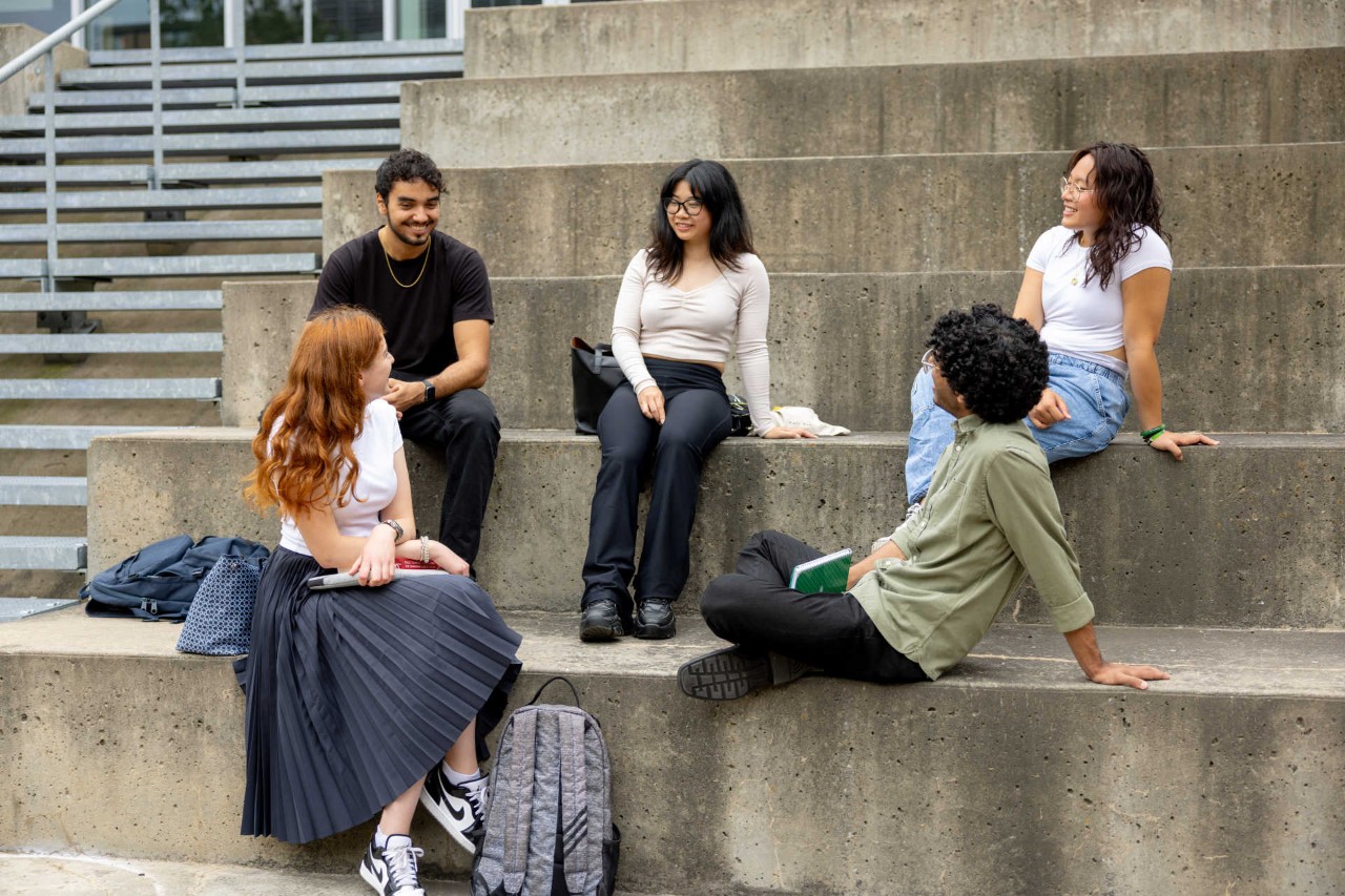 Five students are sitting on large steps and chatting in a casual setting