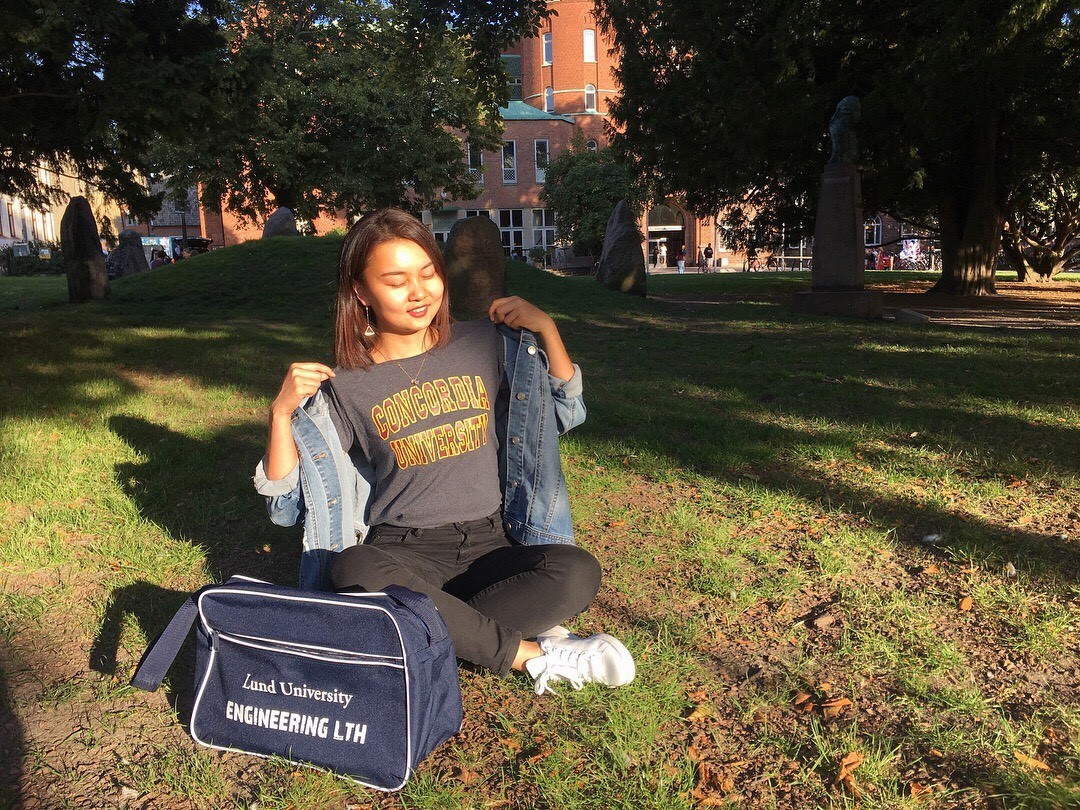 Young woman wearing grey shirt with yellow Concordia University logo, sitting on the ground in front of brick building and trees