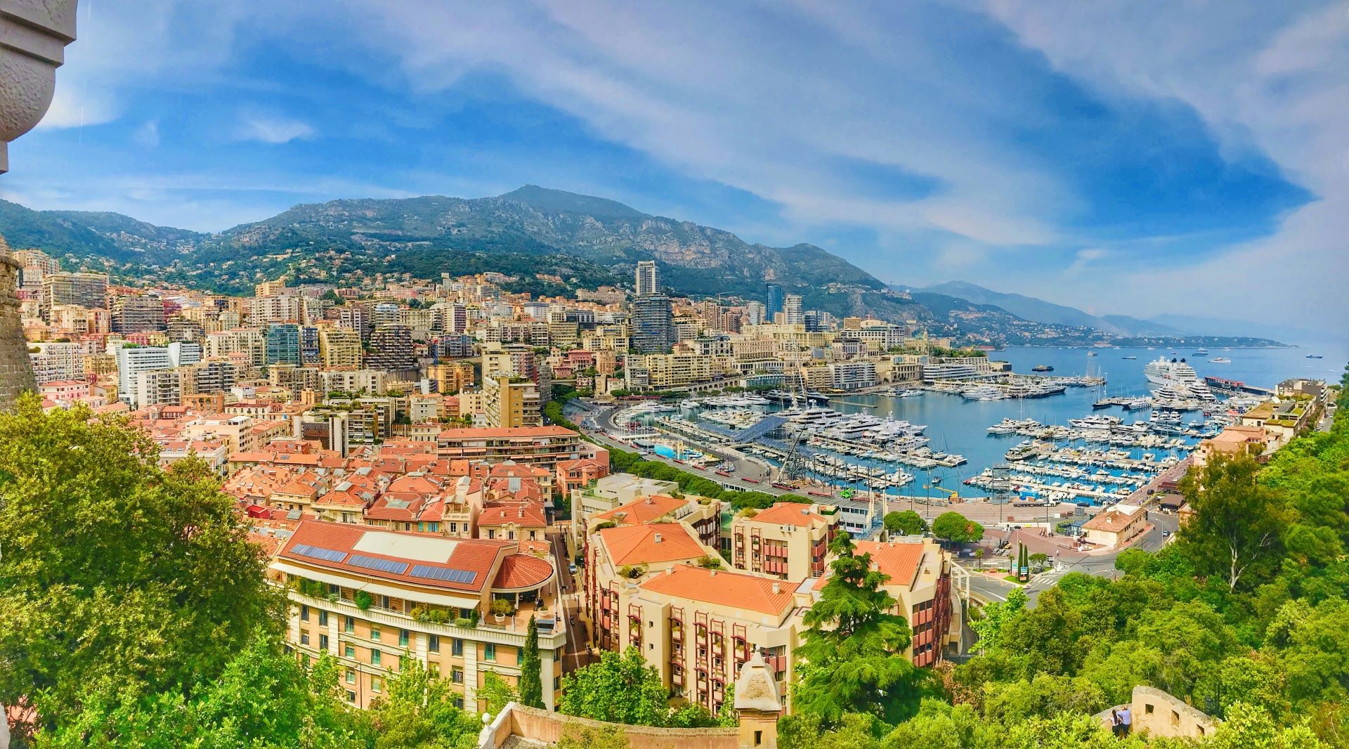 View of Monaco from afar, with orange-roofed buildings, mountains and a bay full of small boats