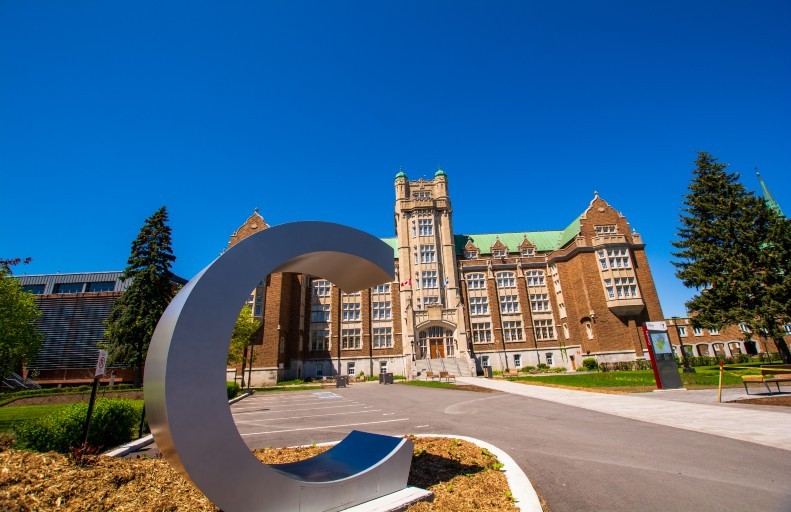 A large "C" sculpture in front of the Administration (AD) Building on the Loyola Campus, surrounded by trees on a clear day.