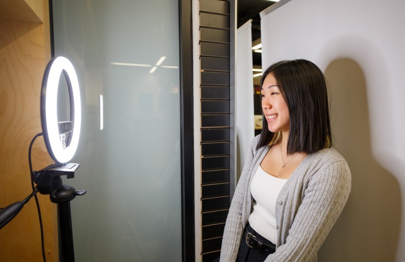 A woman stands in front of a ring light and camera setup, smiling, in an indoor setting with light-colored walls and wooden accents. 