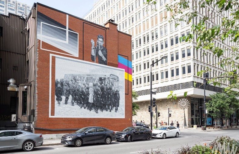 Street scene featuring a mural on a brick building, depicting historical figures and events in black and white, with colorful accents. In the foreground, cars are parked or driving by.