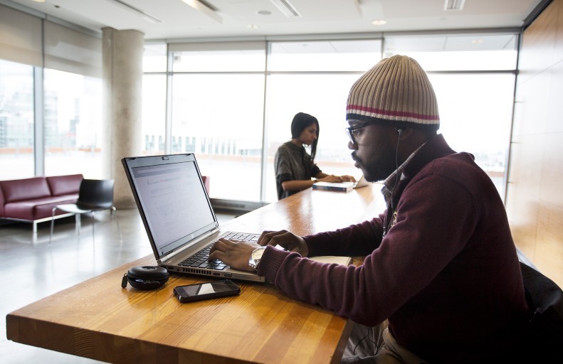 Two people are working on laptops at a wooden table in a modern, bright room with large windows. One person is wearing a knit hat and earphones. A smartphone and a cord are on the table.