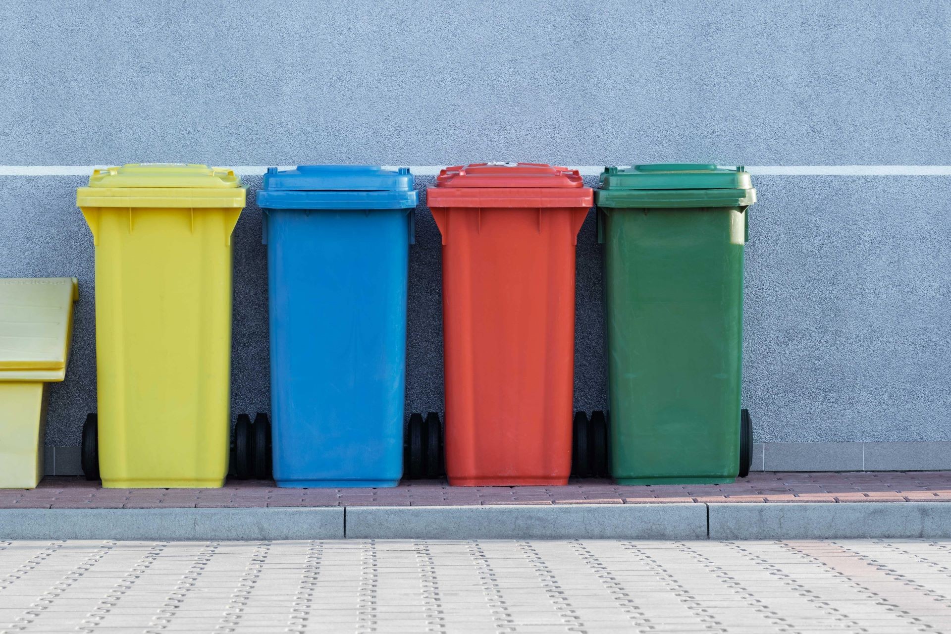 A set of four collection bins of different colours sit on the sidewalk against a building