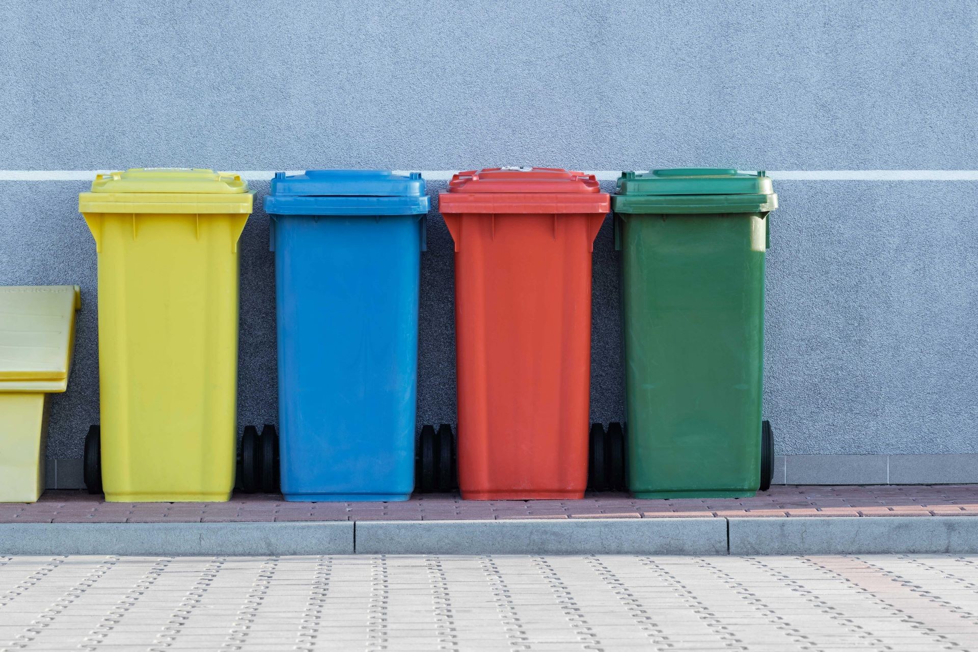 A set of four collection bins of different colours sit on the sidewalk against a building