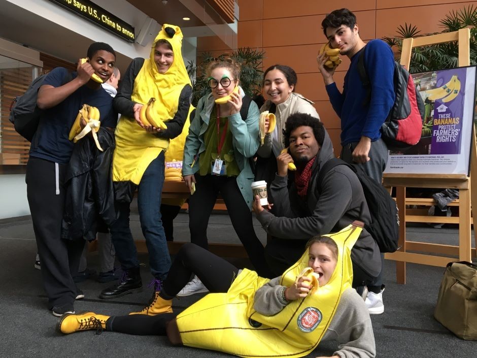 A group of seven students pose in the MB building. Six students are standing, while one is lying on their side. Two students are wearing banana costimes and the others are holding bananas.