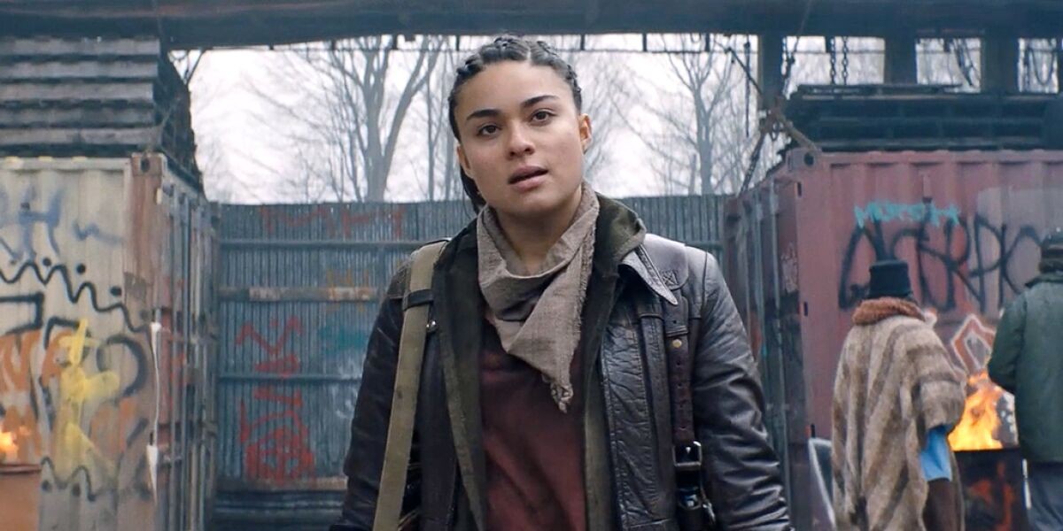 A woman in leather jacket with neck kerchief stands in front of dilapitated and vandalized walls with a defiant expression.