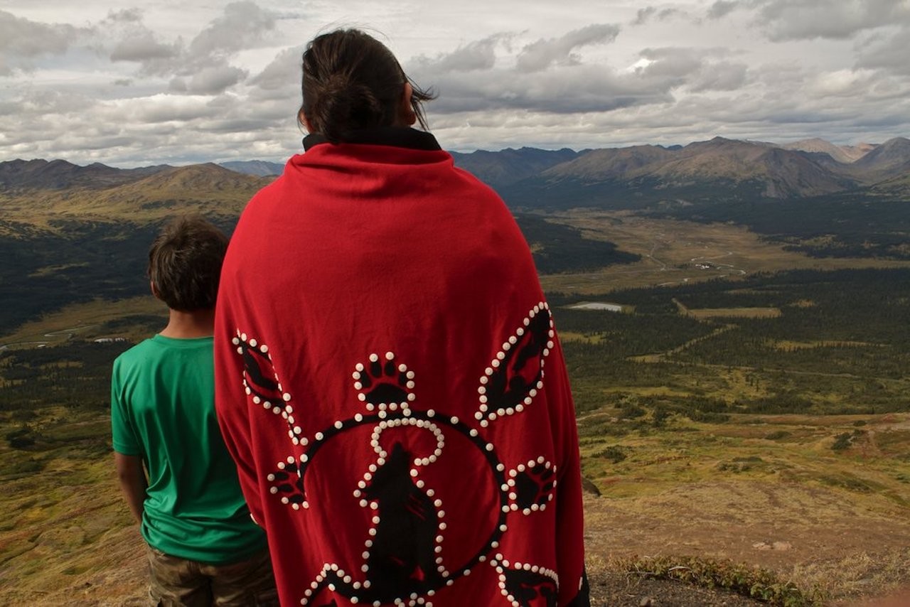 A man in a red cape with beaded designs stands next to a young boy. Both have their backs turned to the camera and are looking out over a mountainous landscape and valley.