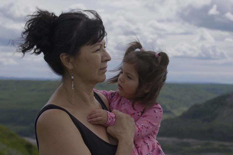 A woman, standing at a high elevation, holds a young girl and looks out at the cloudy sky and landscape while the wind blows in thair hair.