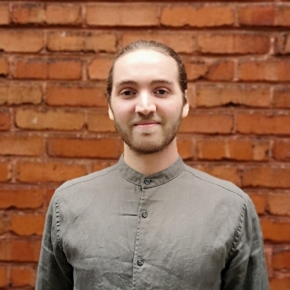 Nicholas Bailey smiles with closed mouth in front of brick wall and wears gray collarless button-up shirt