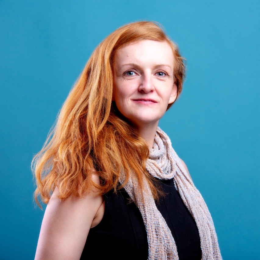 Sara Kosmowska is wearing a black sleeveless top with a pale lavender scarf. She has long red hair and is posed in front of a blue background.
