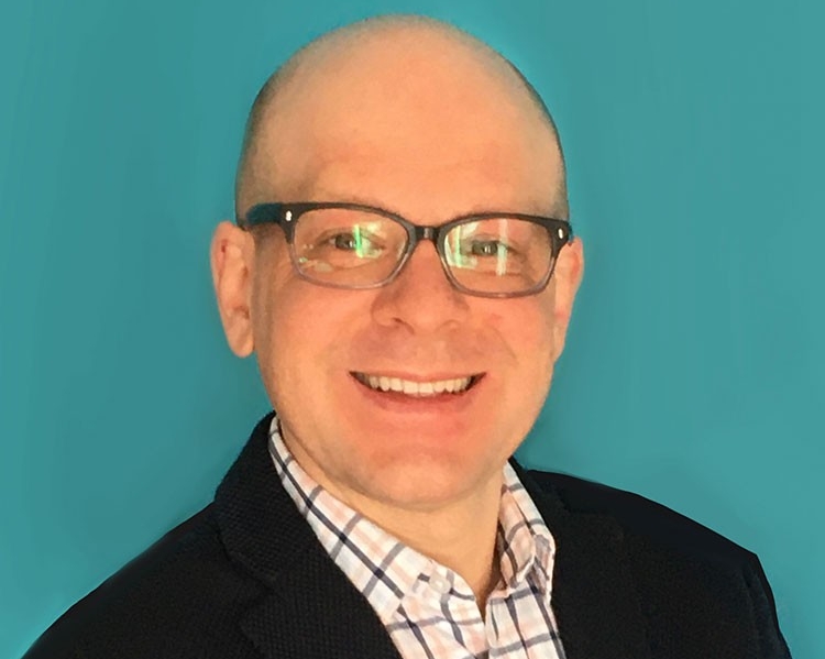 Professor Trofinovich poses in front of a blue background. He is wearing a black jacket and a checkered shirt, with dark-rimmed eyeglasses