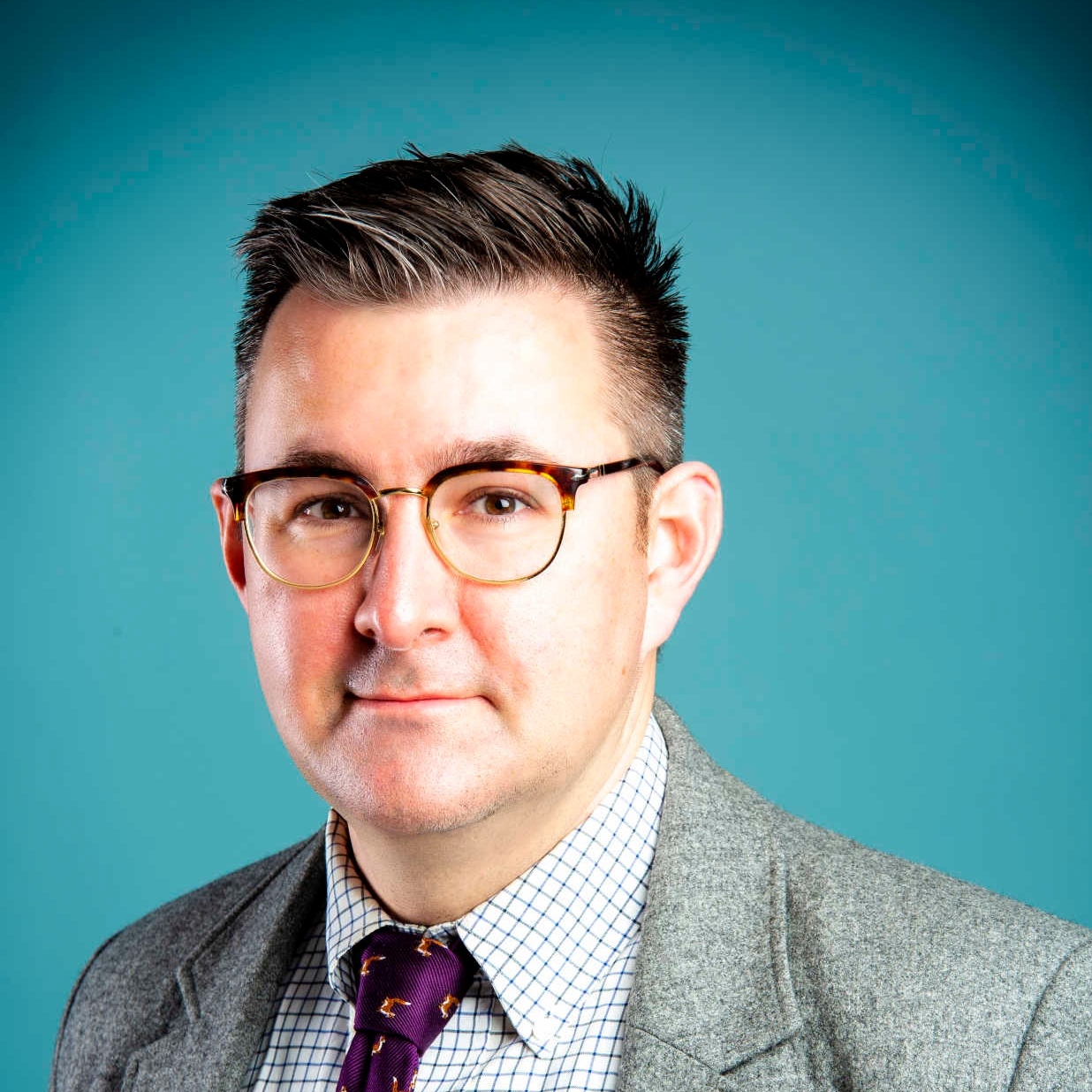 Fenwick McKelvey poses in front of a blue background. He is wearing a grey jacket, a blue-and-white checked shirt, a purple tie, and eyeglasses