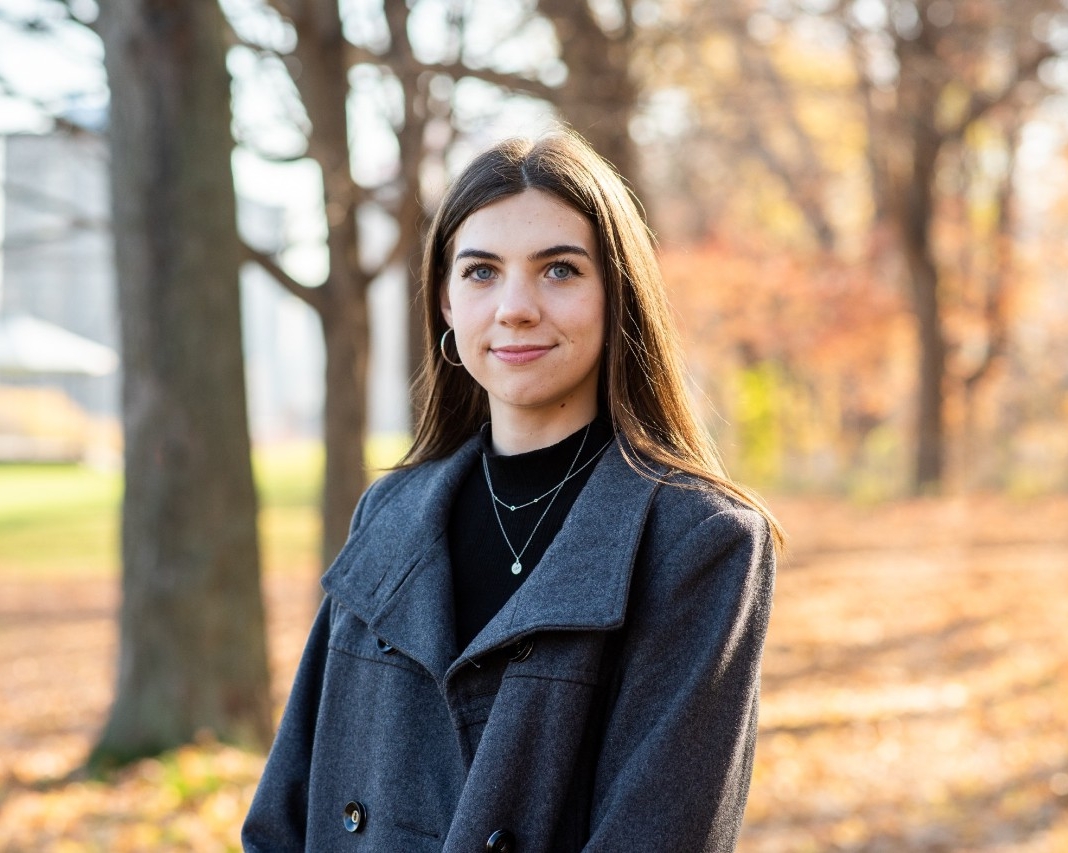 Chloe Cull is poised in front of trees on a sunny day. She is wearing a blue coat and a black turtle neck.