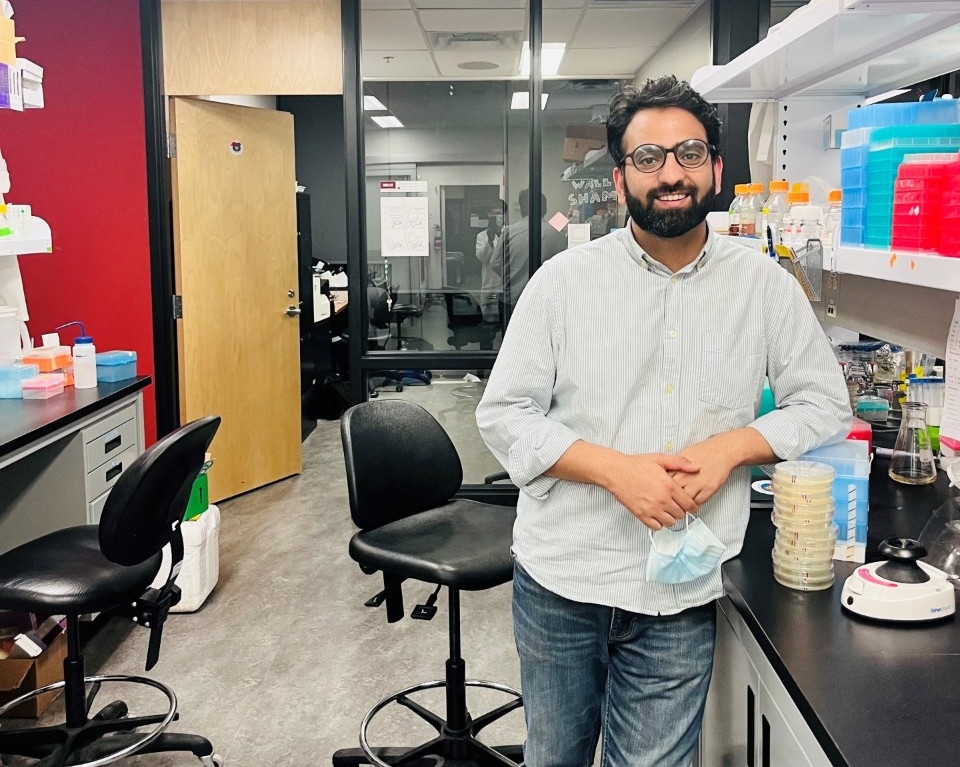 Mudabir Abdullah  poses in the lab. He is leaning against a table, wearing blue jeans and a white button-down shirt