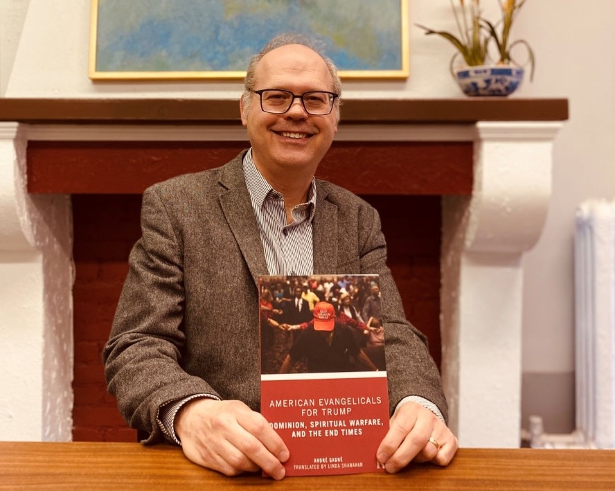 Professor André Gagné sitting at his desk, smiling, holding his book up