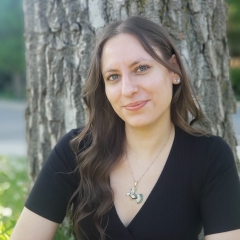 A smiling young woman, with brown hair, wearing a black v-neck top, in front of a tree.