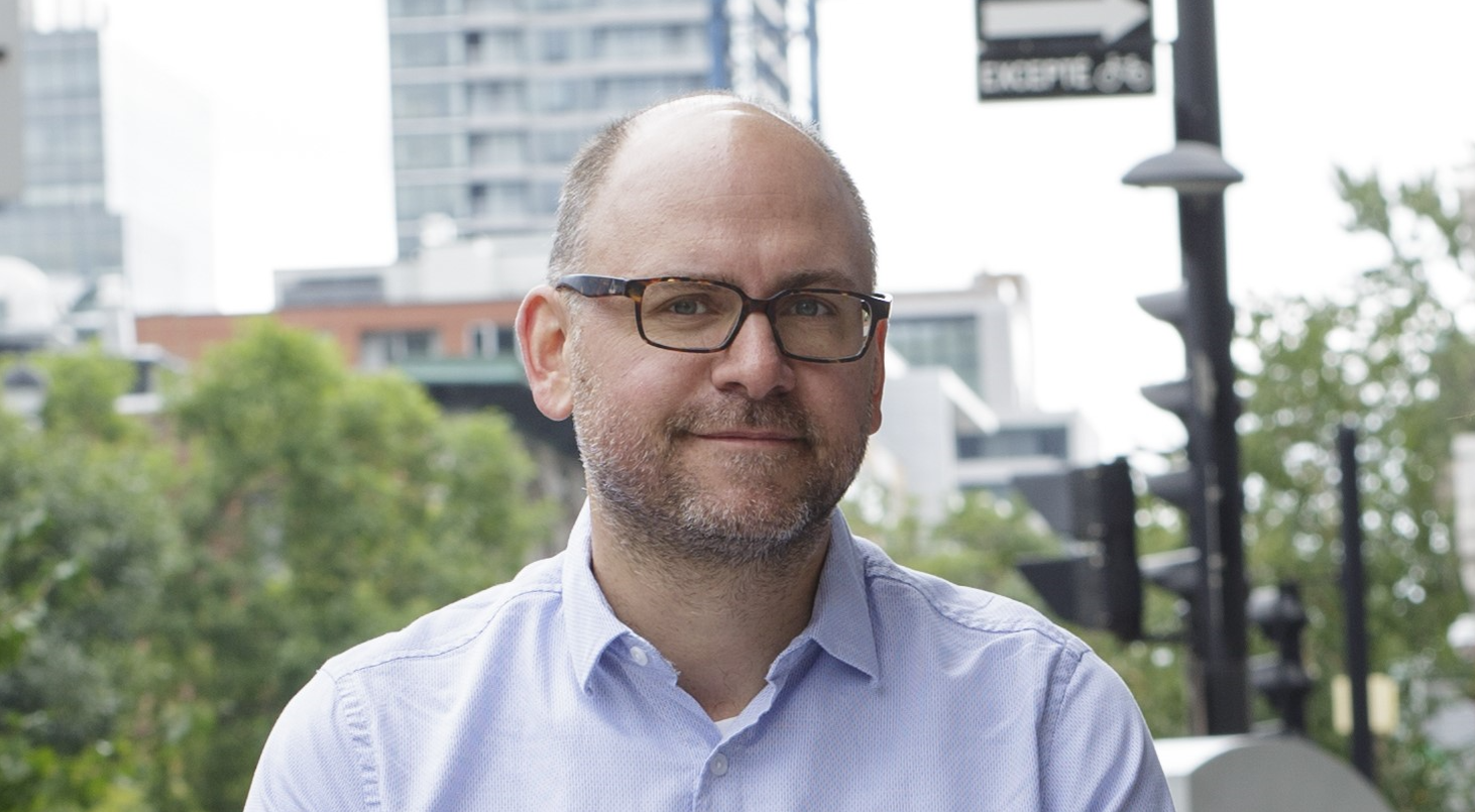 A smiling middle-aged man, wearing glasses and a pale blue shirt, in a city outdoor setting