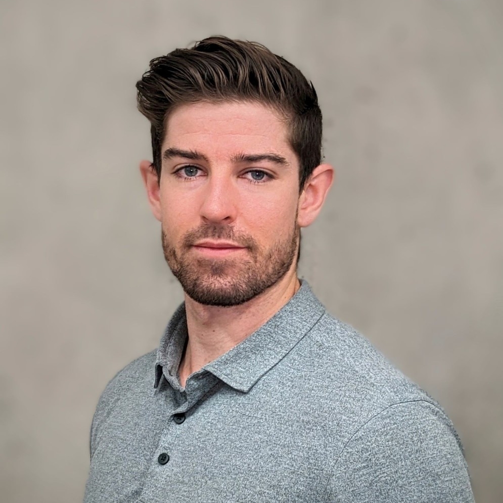 A young man with short brown hair, a moustache and a beard, wearing a grey polo shirt, posing in front of a beige background