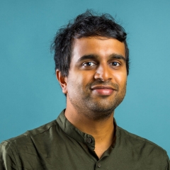 A smiling man with short brown hair, wearing a green top, in front of a turquoise background