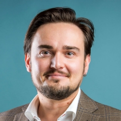 A smiling man with short brown hair, a short beard and moustache, wearing a white collar shirt and a bown suit jacket, with a turquoise background