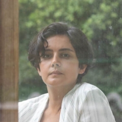 A woman with short brown hair, a white jacket, sitting on the other side of a window, with trees behind her