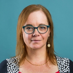 A smiling woman with middle-length blond hair, turquoise glasses and a colourful top, on a turquoise background