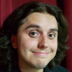 A smiling individual with middle-length brown hair, wearing a black shirt, with a burgundy background.