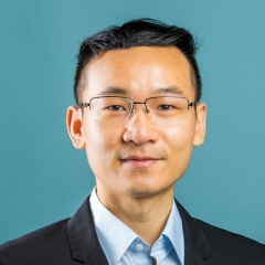 A smiling man with short dark hair and thin glasses, wearing a blue collar shirt and a dark suit jacket, on a turquoise background