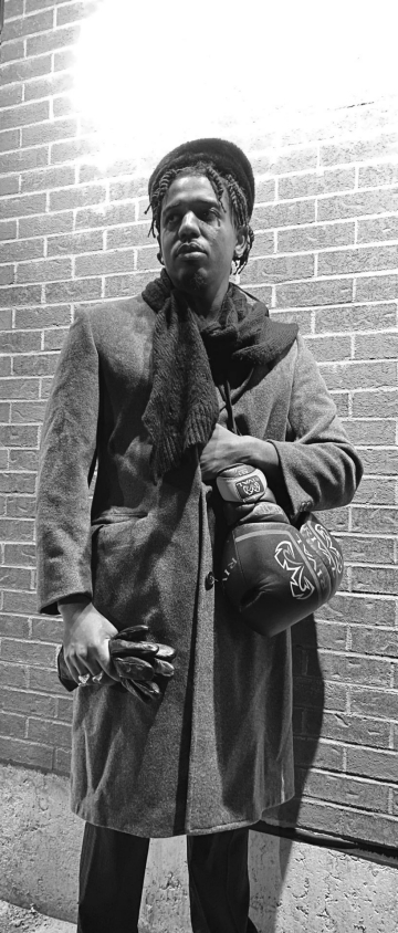 A Black man, impersonating Jack Johnson, the first black heavyweight boxer to win a championship title. He is wearing a wool coat and hat, leather gloves, gold jewelry, a bag, and is standing in front of a brick wall. Photo is black and white.