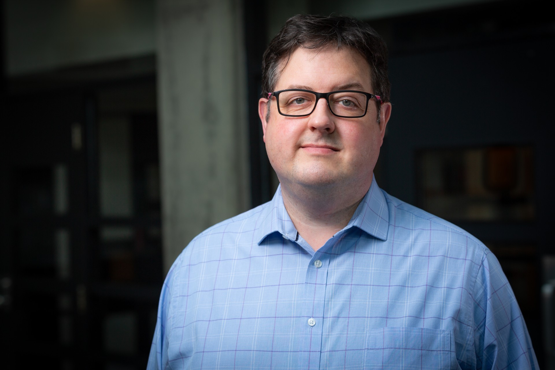 A man with short brown hair and glasses, wearing a blue collar shirt