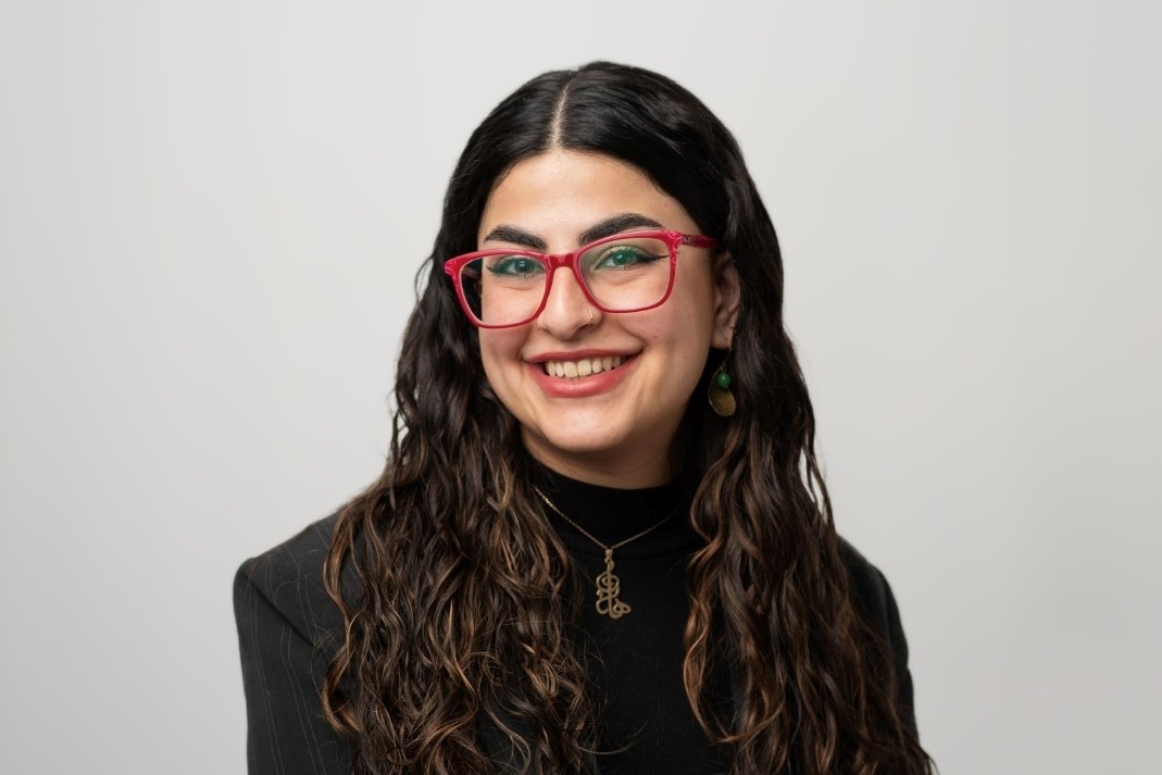 A smiling young woman with long dark hair, wearing red glasses, and a black suit jacket