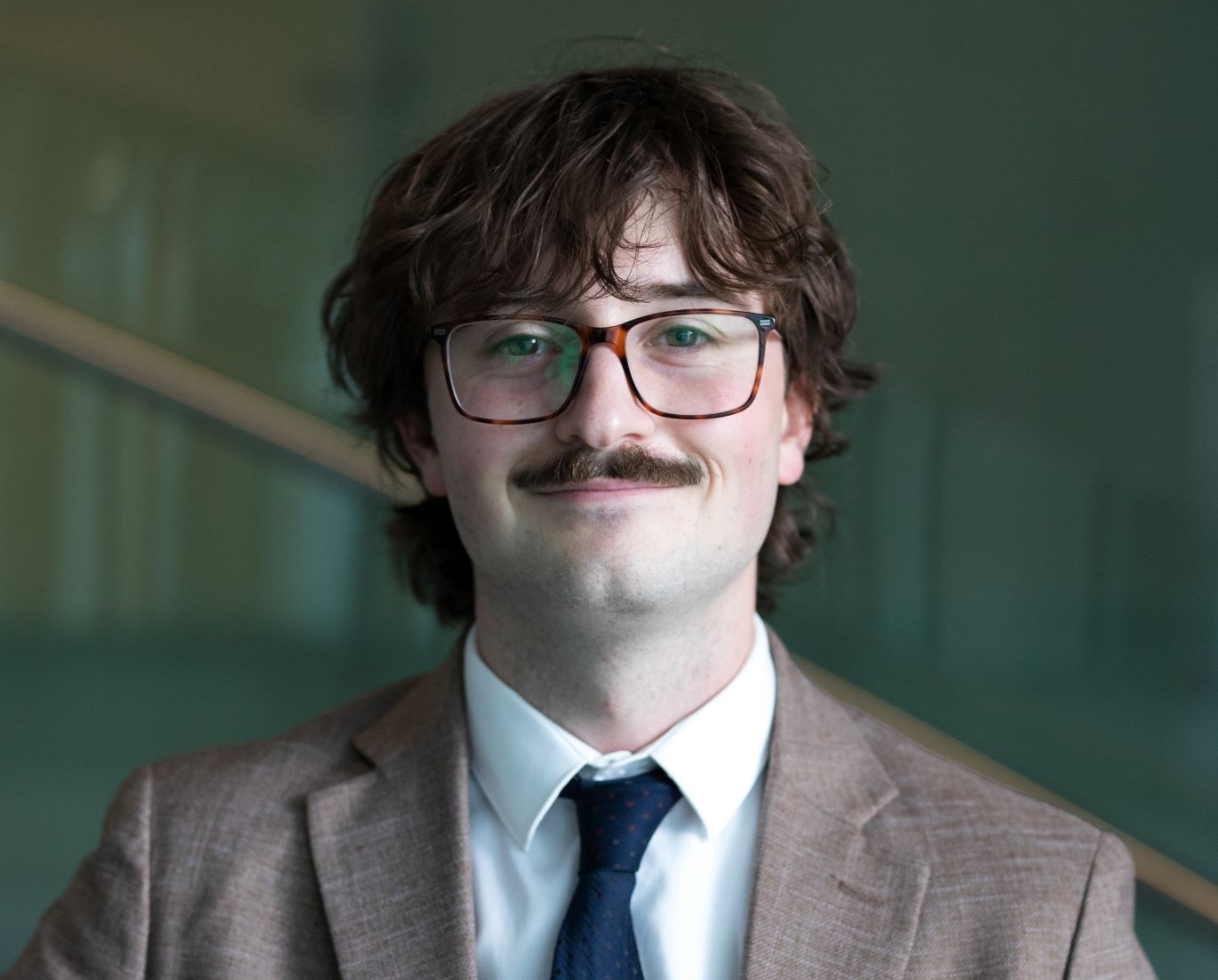 A smiling young man with mid-length brown curly hair and a moustache, wearing glasses, a white collar shirt, a beige suit jacket and a navy blue tie