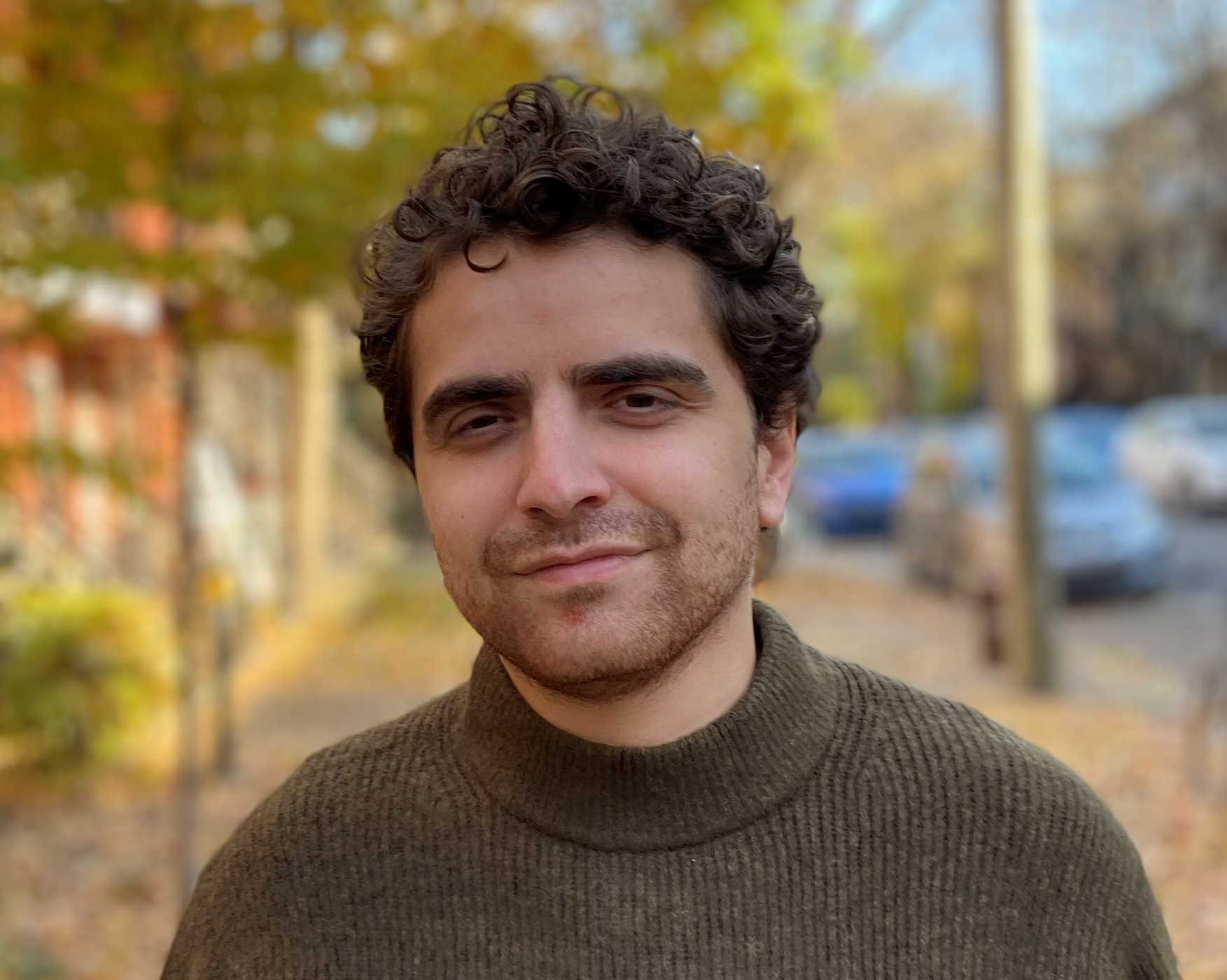 A smiling man with brown short curly hair, wearing a brown turtleneck, standing outside in front of trees