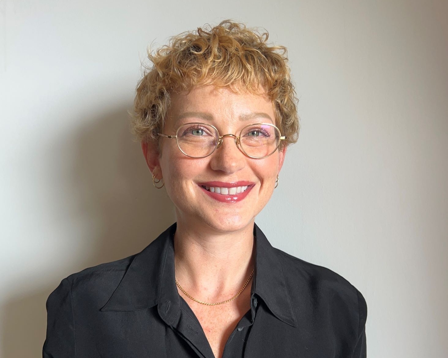 A smiling young caucasian woman with short blond hair, a black suit, and glasses, standing in front of a grey wall